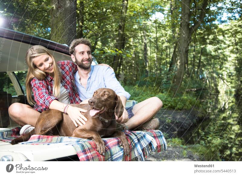 Lächelndes junges Paar mit Hund sitzt im Auto an einem Bach im Wald Hunde lächeln Wagen PKWs Automobil Autos Forst Wälder Bäche Baeche Pärchen Paare
