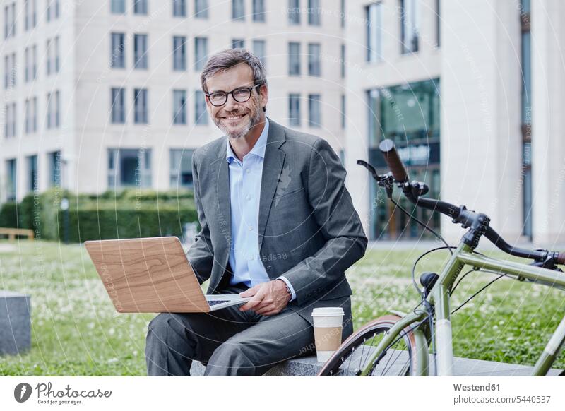 Porträt eines lächelnden reifen Geschäftsmannes im Freien mit Laptop, Kaffee zum Mitnehmen und Fahrrad Notebook Laptops Notebooks Businessmann Businessmänner