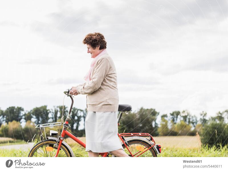 Ältere Frau schiebt Fahrrad in ländlicher Landschaft Bikes Fahrräder Räder Rad lächeln schieben anschieben Seniorin älter Seniorinnen alt Raeder Senioren