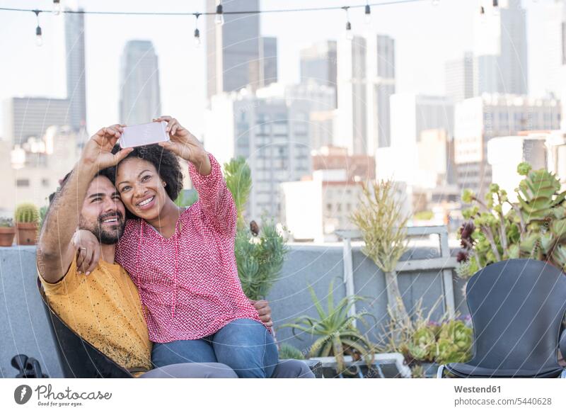 Ein Paar sitzt auf einer Dachterrasse und macht Selfies mit dem Smartphone Pärchen Paare Partnerschaft fotografieren sitzen sitzend lächeln Dachterrassen Mensch