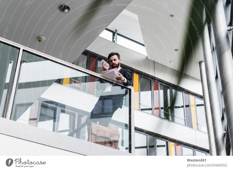 Geschäftsmann, der auf dem Treppenabsatz eines Bürogebäudes steht, telefoniert, während er Dokumente liest Europäer Kaukasier Europäisch kaukasisch Geländer