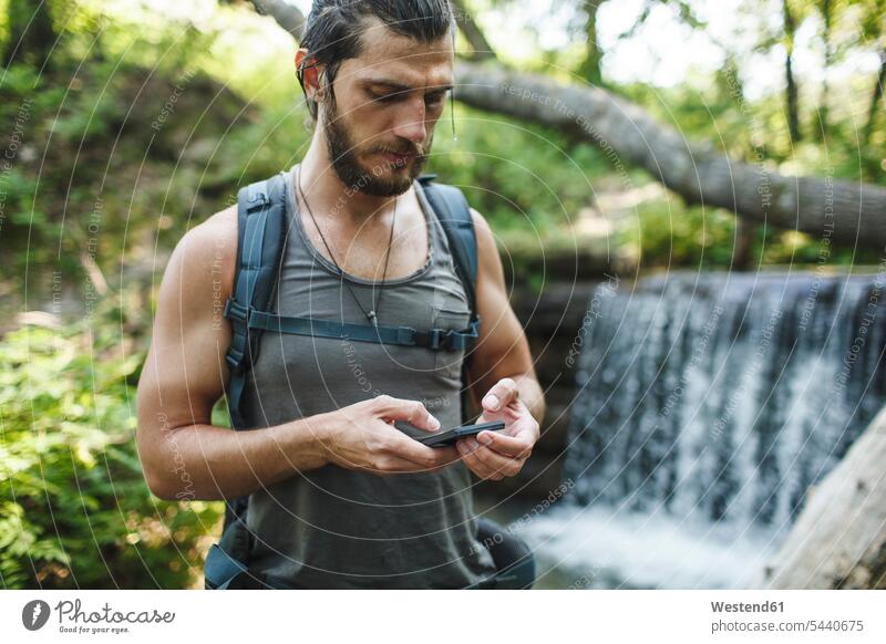 Junger Mann hält Handy an einem Wasserfall im Wald wandern Wanderung Mobiltelefon Handies Handys Mobiltelefone Männer männlich Wanderer Telefon telefonieren
