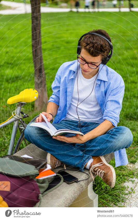 Junger Mann mit Rennrad und Kopfhörer sitzt auf einer Bank und liest ein Buch Kopfhoerer Männer männlich Bücher Erwachsener erwachsen Mensch Menschen Leute