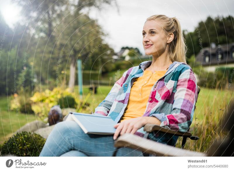 Lächelnde junge Frau mit Buch entspannt im Garten Gärten Gaerten Gartenanlage Gartenanlagen Bücher weiblich Frauen entspanntheit relaxt Erwachsener erwachsen