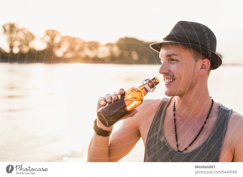 Porträt eines jungen Mannes mit Hut am Flussufer entspannt entspanntheit relaxt lächeln Männer männlich Bier Entspannung relaxen entspannen Erwachsener