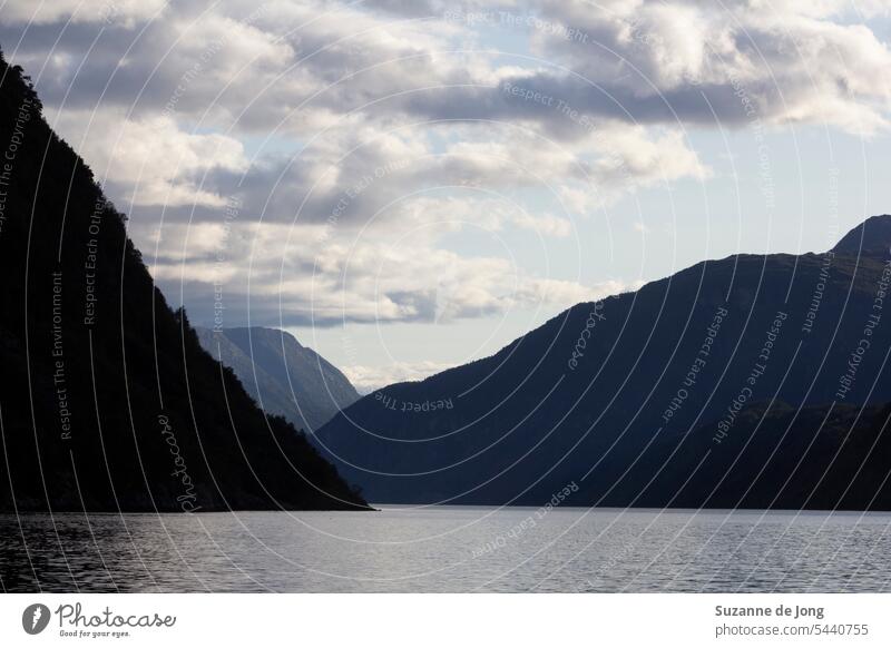 Malerischer Blick auf einen Fjord und Berge in Norwegen an einem schönen Sommernachmittag, mit blauem Himmel und einigen Wolken. Das Licht hinter dem Berg schafft eine schöne Silhouette. Die Stimmung des Bildes ist beruhigend.