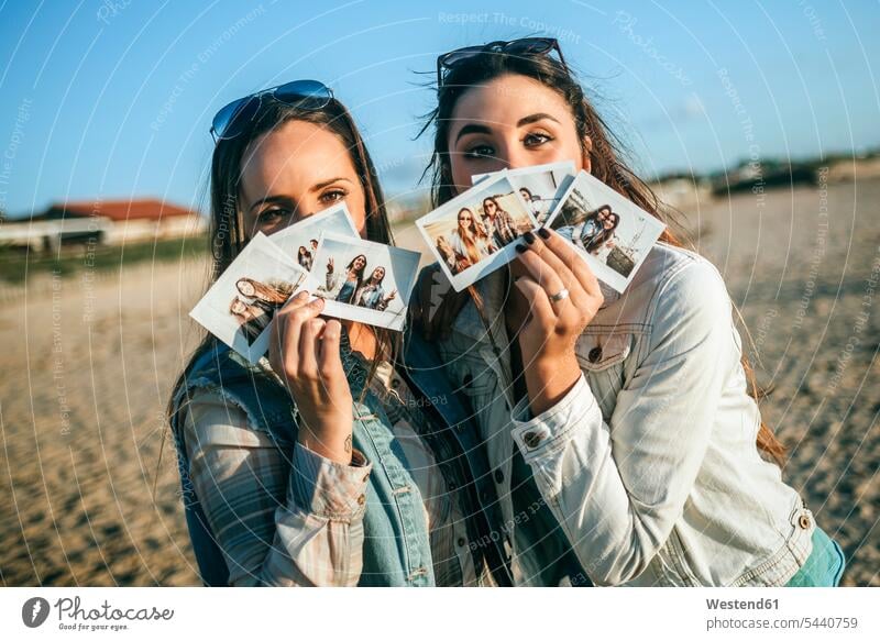 Zwei junge Frauen halten sich Sofortfotos vor das Gesicht Freundinnen Strand Beach Straende Strände Beaches Sofortbild Polaroids Sofortbilder Freunde