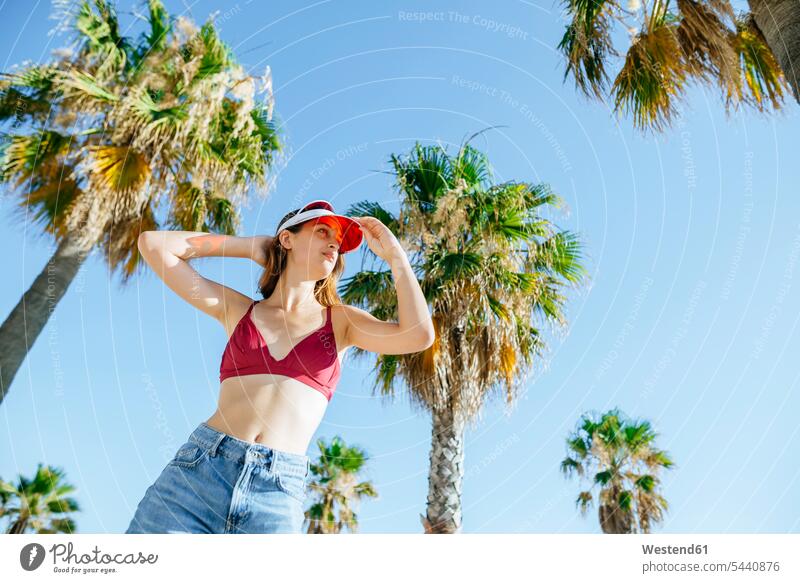 Bildnis einer jungen Frau mit Bikini und Sonnenblende vor blauem Himmelshintergrund und Palmen stehen stehend steht weiblich Frauen Erwachsener erwachsen Mensch