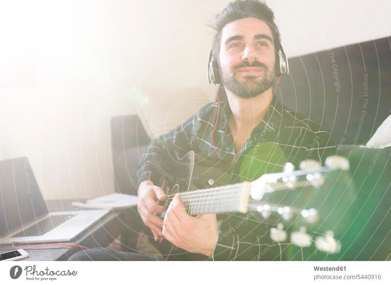 Junger Mann spielt zu Hause Gitarre und trägt Kopfhörer, die an einen Laptop angeschlossen sind Gitarren Notebook Laptops Notebooks lächeln Männer männlich