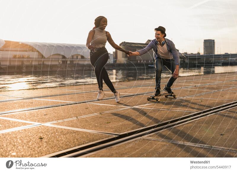 Junges Paar beim Skateboarden am Flussufer jung multikulturell ziehen Hände halten fröhlich aktiv Freizeitsport Freizeitaktivität Hand in Hand Freude Mensch