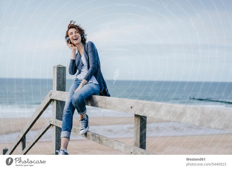 Glückliche Frau sitzt am Strand auf einem Geländer und hört Musik Beach Straende Strände Beaches lachen Kopfhörer Kopfhoerer weiblich Frauen positiv Emotion