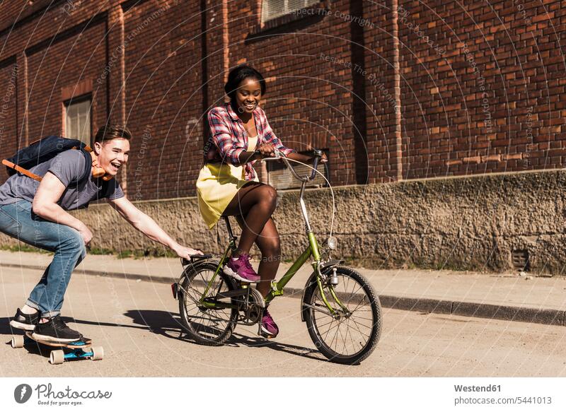 Junge Frau auf Fahrrad ziehender junger Mann, stehend auf Skateboard multikulturell Skateboarden Skateboardfahren Skateboarding fröhlich Fröhlichkeit Frohsinn