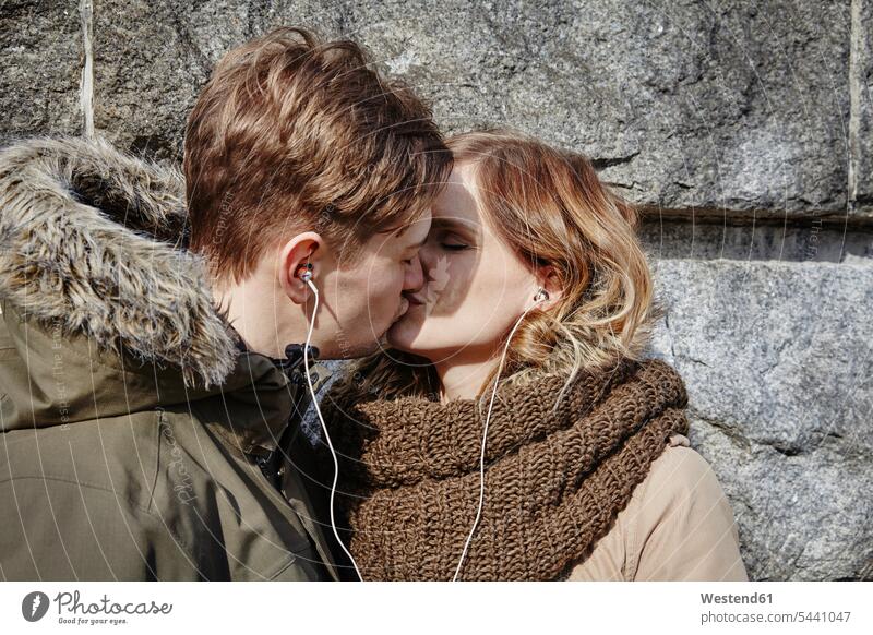 Junges Paar mit gemeinsamen Ohrstöpseln küsst sich an der Steinmauer küssen Küsse Kuss Pärchen Paare Partnerschaft Ohrhörer Mensch Menschen Leute People