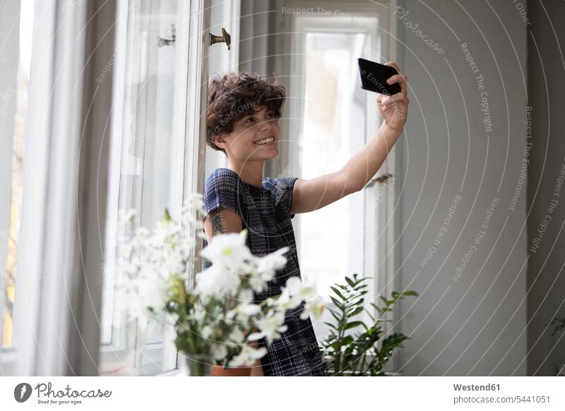 Lächelnde junge Frau steht vor einem Fenster und macht ein Selfie mit ihrem Smartphone Selfies fotografieren weiblich Frauen Erwachsener erwachsen Mensch