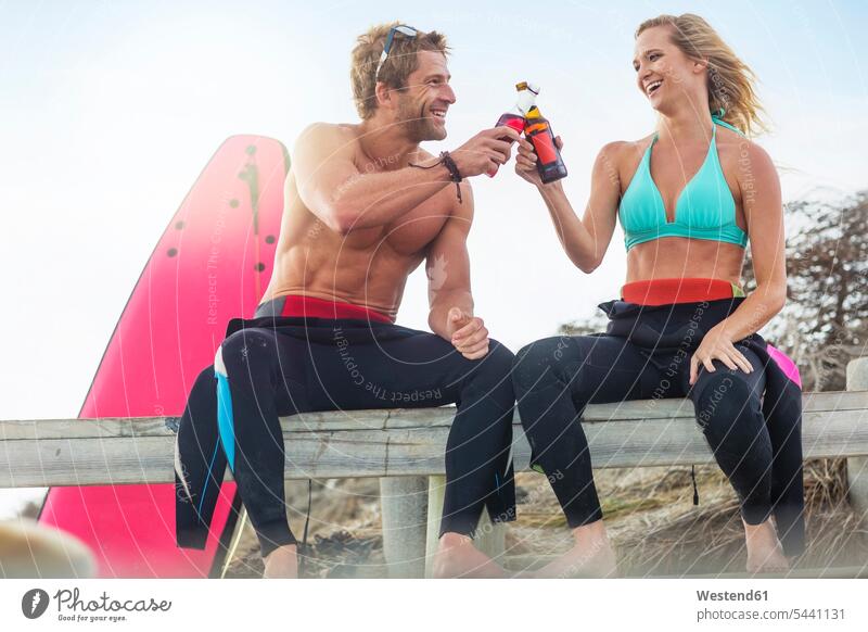 Glückliches Paar mit Surfbrett bei einem Drink am Strand lachen trinken Beach Straende Strände Beaches Pärchen Paare Partnerschaft positiv Emotion Gefühl