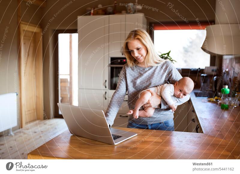 Lächelnde Mutter mit Baby in der Küche mit Laptop Notebook Laptops Notebooks Babies Babys Säuglinge Kind Kinder Mami Mutti Mütter Mama Computer Rechner Mensch