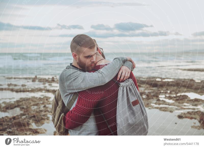Junges schwules Paar mit Rucksäcken umarmt sich am Strand Pärchen Paare Partnerschaft Beach Straende Strände Beaches Mensch Menschen Leute People Personen