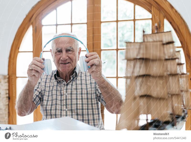 Porträt eines selbstbewussten älteren Mannes beim Auspacken der Kopfhörer aus dem Paket mit dem Schiffsmodell auf dem Tisch Modellboot auspacken lächeln