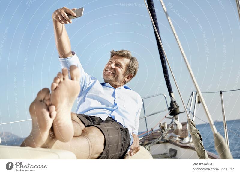 Lächelnder reifer Mann, der auf dem Deck seines Segelbootes liegt und ein Selfie mit seinem Handy macht Segeln segelnd segelt Selfies Männer männlich Bootsport