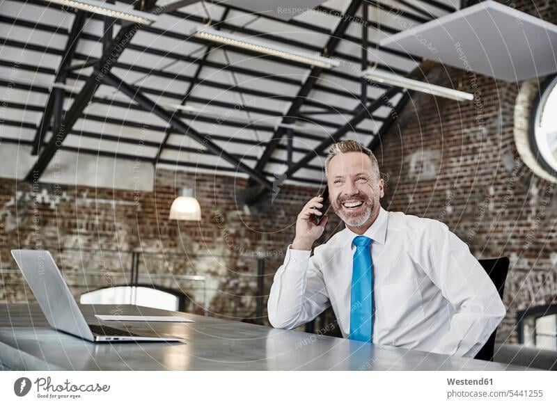 Glücklicher Geschäftsmann am Handy sitzt am Tisch in einem Loft telefonieren anrufen Anruf telephonieren sitzen sitzend Mobiltelefon Handies Handys
