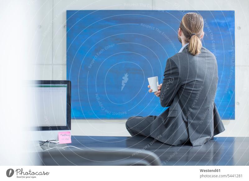 Geschäftsmann im Büro sitzt auf dem Schreibtisch und betrachtet ein blaues Gemälde sitzen sitzend Bild Gemaelde Bilder Businessmann Businessmänner