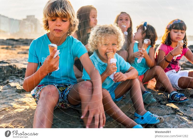 Eine Gruppe von sechs Kindern isst Eiscreme am Strand Kids Speiseeis sitzen sitzend sitzt essen essend Mensch Menschen Leute People Personen Süßspeise Süsses