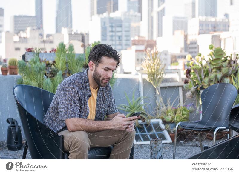 Junger Mann sitzt auf der Dachterrasse und hört Smartphone-Nachrichten ab Männer männlich Stuhl Stuehle Stühle Handy Mobiltelefon Handies Handys Mobiltelefone