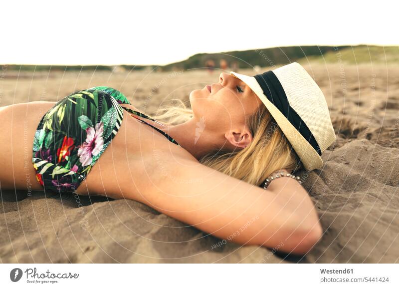 Junge Frau mit Hut, die mit geschlossenen Augen am Strand liegt weiblich Frauen entspannt entspanntheit relaxt Beach Straende Strände Beaches liegen liegend