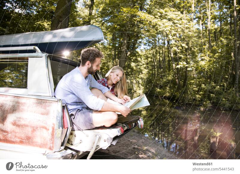 Lächelndes junges Paar mit Karte und Kanu im Auto an einem Bach Landkarte Landkarten Bäche Baeche Wald Forst Wälder Pärchen Paare Partnerschaft Kanus Wagen PKWs