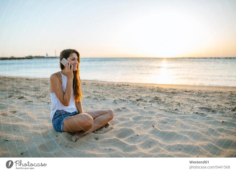 Junge Frau telefoniert bei Sonnenuntergang am Strand mit ihrem Handy Mobiltelefon Handies Handys Mobiltelefone Beach Straende Strände Beaches Meer Meere