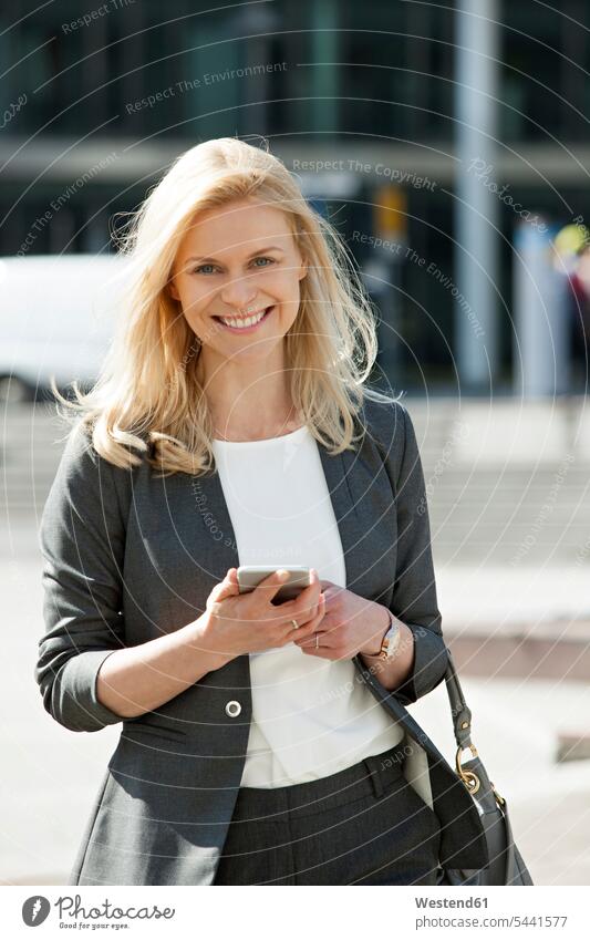 Porträt einer lächelnden blonden Geschäftsfrau mit Mobiltelefon und Handtasche Geschäftsfrauen Businesswomen Businessfrauen Businesswoman Portrait Porträts
