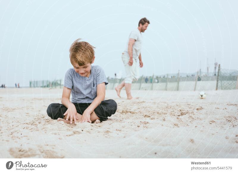 Lächelnder kleiner Junge sitzt auf dem Sand am Strand, während sein Vater im Hintergrund mit dem Ball spielt Beach Straende Strände Beaches Papas Väter Vati