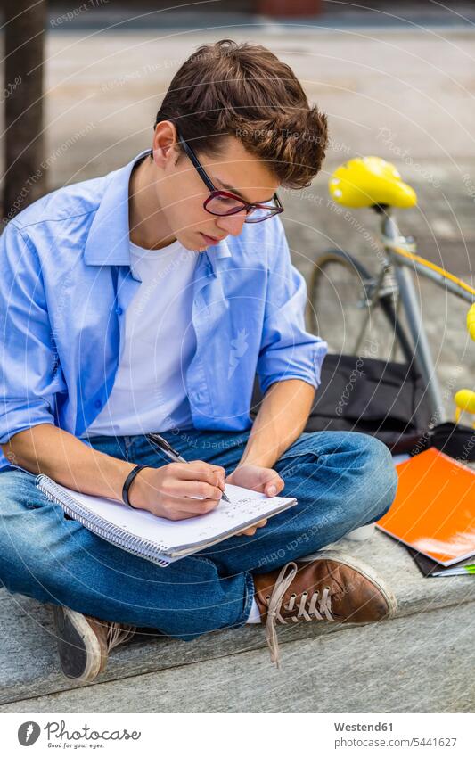 Junger Mann mit Rennrad sitzt auf Bank und schreibt auf Notizblock Student Hochschueler Studierender Hochschüler Studenten lernen schreiben aufschreiben