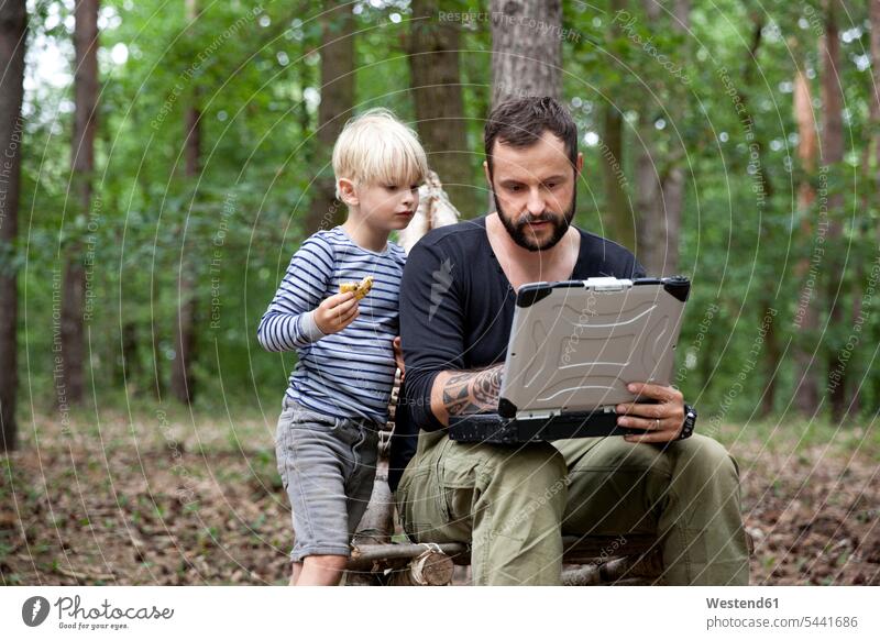 Vater und Sohn sitzen mit Laptop auf selbstgebautem Holzstuhl im Wald Notebook Laptops Notebooks Mann Männer männlich Söhne Forst Wälder Junge Buben Knabe