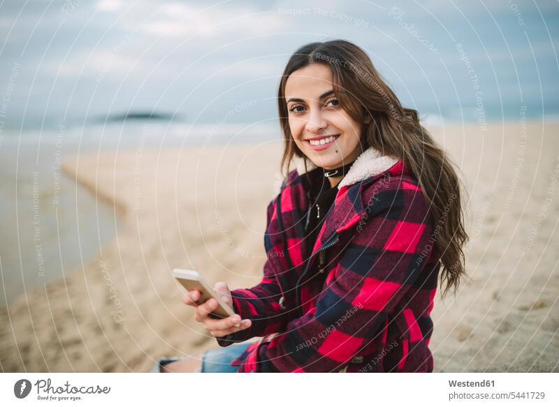 Porträt einer lächelnden jungen Frau mit Smartphone am Strand Beach Straende Strände Beaches weiblich Frauen Portrait Porträts Portraits Erwachsener erwachsen