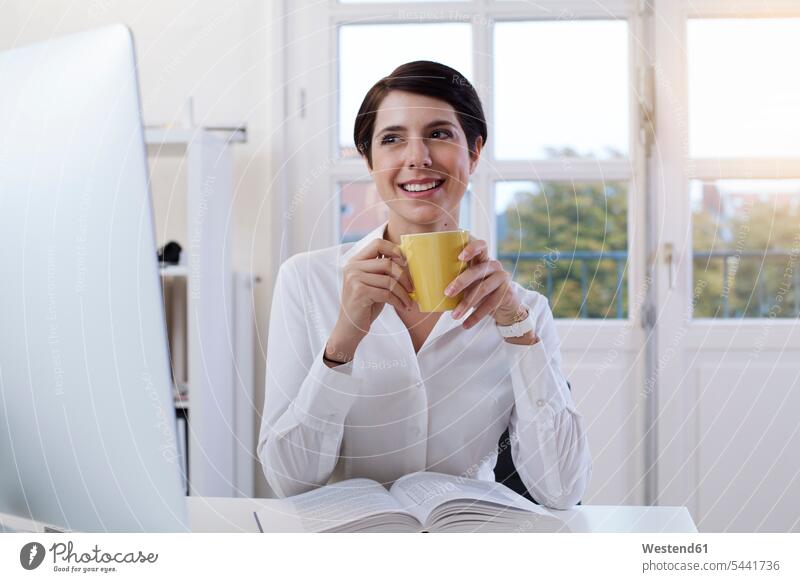 Lächelnde Frau hält Tasse Kaffee am Schreibtisch im Büro Office Büros weiblich Frauen lächeln Arbeitsplatz Arbeitsstätte Arbeitstelle Erwachsener erwachsen