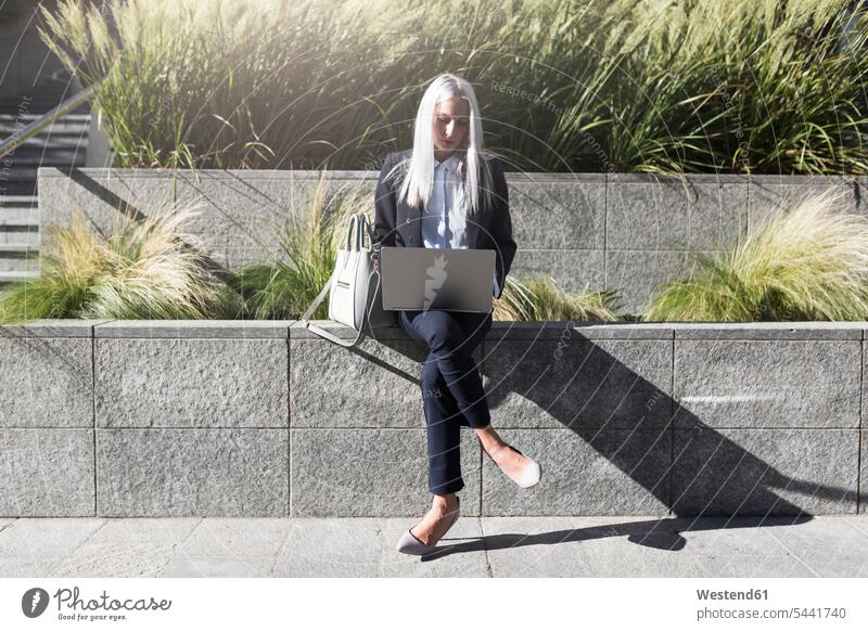 Junge Geschäftsfrau, die in der Stadt mit einem Laptop an einer Wand sitzt Geschäftsfrauen Businesswomen Businessfrauen Businesswoman Notebook Laptops Notebooks