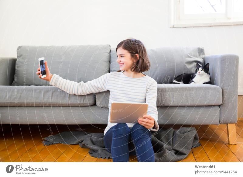 Glückliches Mädchen mit Tablette sitzt auf dem Boden des Wohnzimmers nehmen selfie mit Smartphone sitzen sitzend Selfie Selfies Fußboden Fußboeden Fussboeden