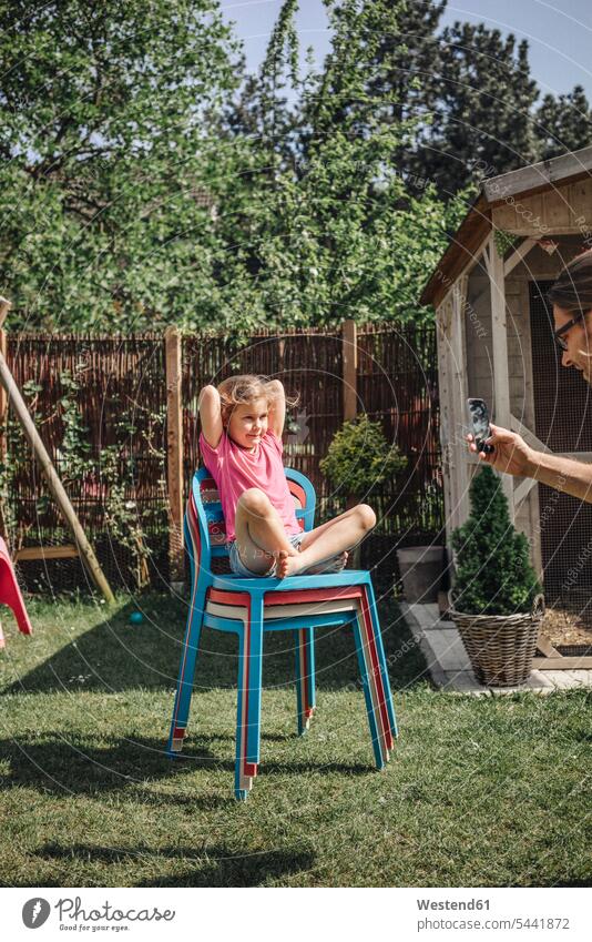 Vater macht Handyfoto von Tochter, die auf einem Stuhl im Garten sitzt Töchter Mobiltelefon Handies Handys Mobiltelefone Gärten Gaerten fotografieren Papas