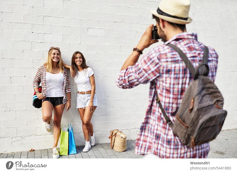 Mann fotografiert zwei junge Frauen, die mit Taschen an einer weißen Wand stehen lächeln Freunde fotografieren stehend steht Freundschaft Kameradschaft Wände