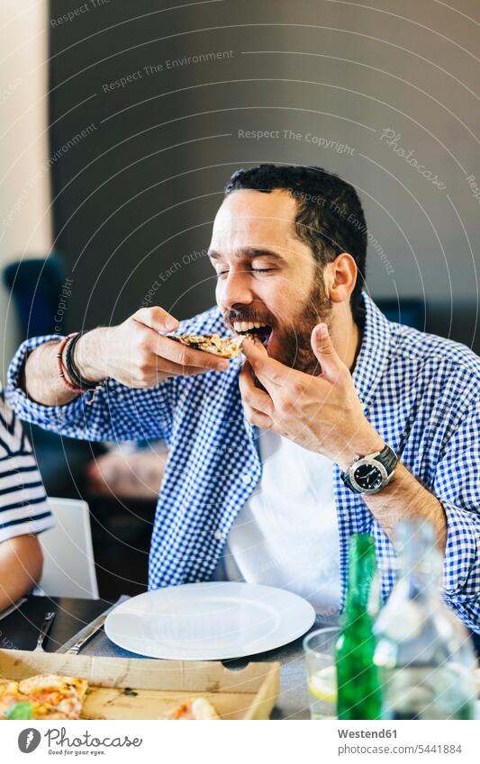 Junger Mann genießt ein Stück Pizza am Esstisch Männer männlich Pizzen essen essend Erwachsener erwachsen Mensch Menschen Leute People Personen Essen Food