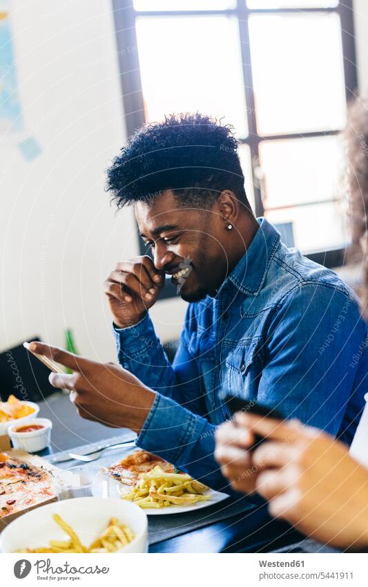 Lächelnder Mann schaut auf Handy am Esstisch Mobiltelefon Handies Handys Mobiltelefone lächeln essen essend Tisch Tische Telefon telefonieren Kommunikation