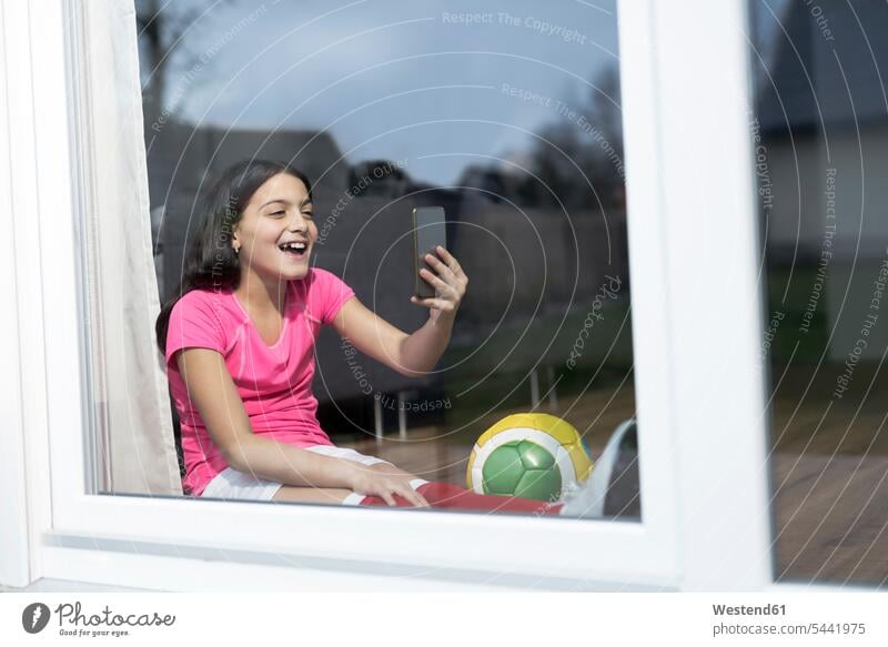 Glückliches Mädchen im Fußball-Outfit sitzt auf dem Boden im Wohnzimmer und macht ein Selfie Fußbälle Fussball Handy Mobiltelefon Handies Handys Mobiltelefone