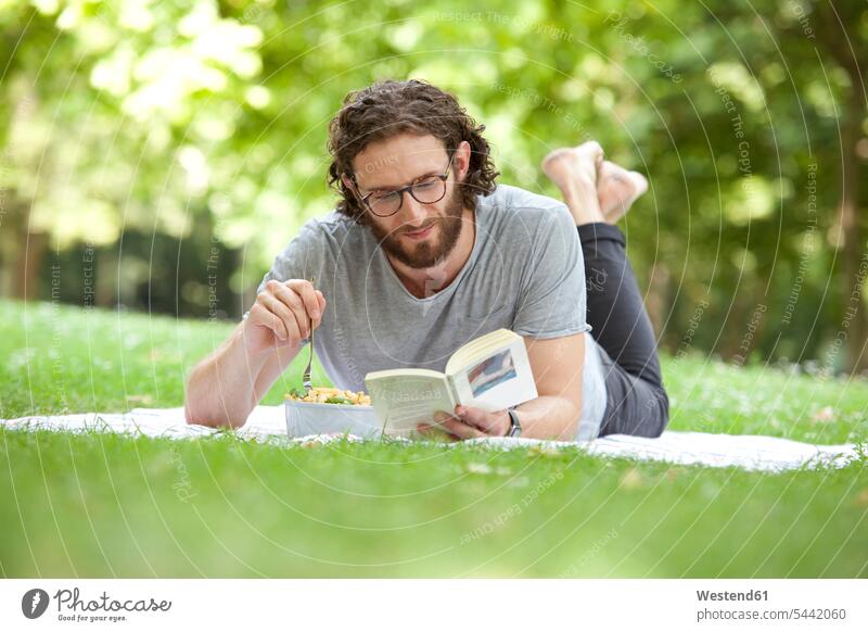 Mann liest Buch auf einer Decke in einem Park, während er Nudelsalat isst Männer männlich Bücher Erwachsener erwachsen Mensch Menschen Leute People Personen