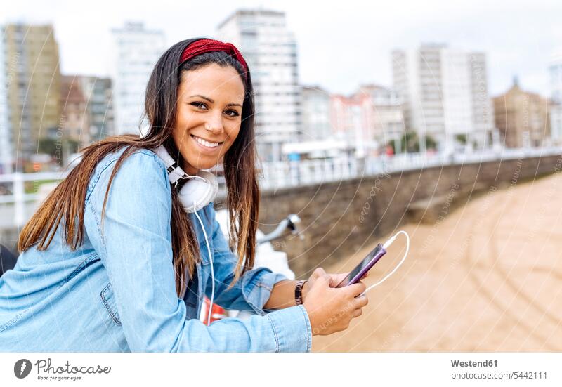 Spanien, Gijon, lächelnde junge Frau mit Handy und Kopfhörern an der Uferpromenade weiblich Frauen Kopfhoerer Mobiltelefon Handies Handys Mobiltelefone