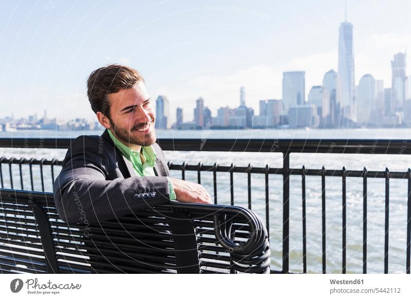 USA, lächelnder Mann sitzt auf einer Bank am Hafen von New Jersey mit Blick auf Manhattan Männer männlich Sitzbänke Bänke Sitzbank sitzen sitzend New York