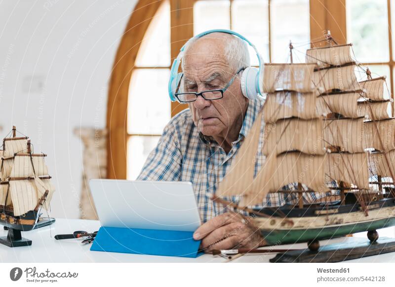 Älterer Mann mit Kopfhörern arbeitet auf Modellschiff auf Tisch mit Tablette Kopfhoerer Senior ältere Männer älterer Mann Senioren männlich Modellboot