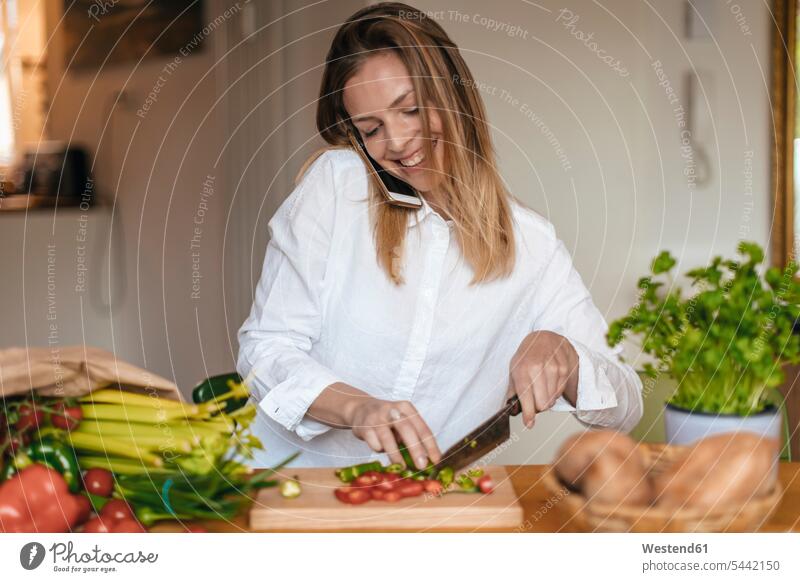 Lächelnde Frau am Telefon, die in der Küche Gemüse schneidet schneiden abschneiden kleinschneiden telefonieren anrufen Anruf telephonieren weiblich Frauen