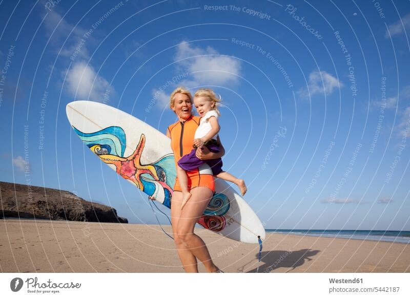 Spanien, Fuerteventura, glückliche Mutter und Tochter spazieren mit Surfbrett am Strand Spaß Spass Späße spassig Spässe spaßig Mami Mutti Mütter Mama Töchter
