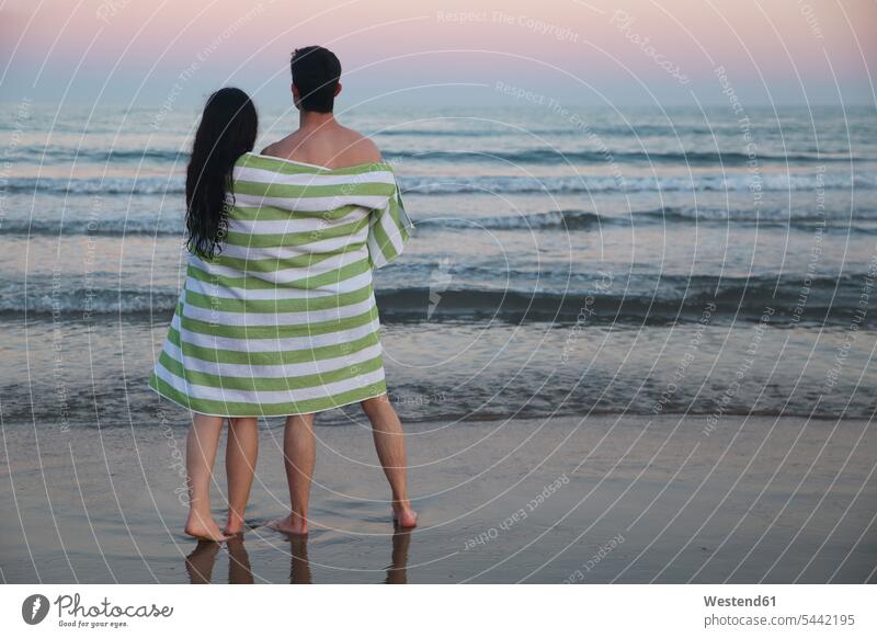 Rückenansicht eines jungen Paares am Strand, das gemeinsam in ein Handtuch gewickelt den Sonnenuntergang betrachtet Pärchen Partnerschaft Sonnenuntergänge Beach